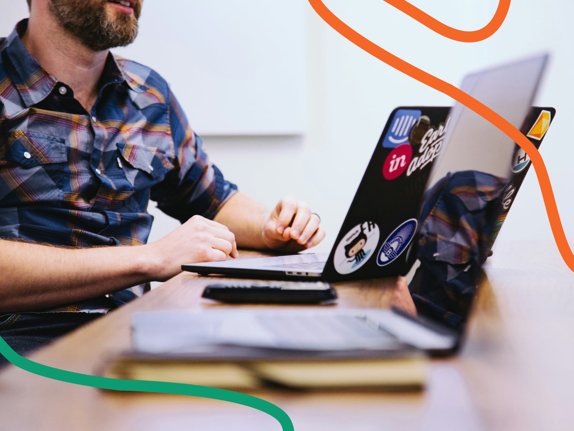 NDS-Kultusministerium_Erzieher-C5-grafik Ein Mann mit Bart und kariertem Hemd sitzt an einem Holztisch und arbeitet an einem Laptop mit mehreren Aufklebern. Im Vordergrund sind ein weiteres Notebook, ein Notizbuch und eine Fernbedienung zu sehen.