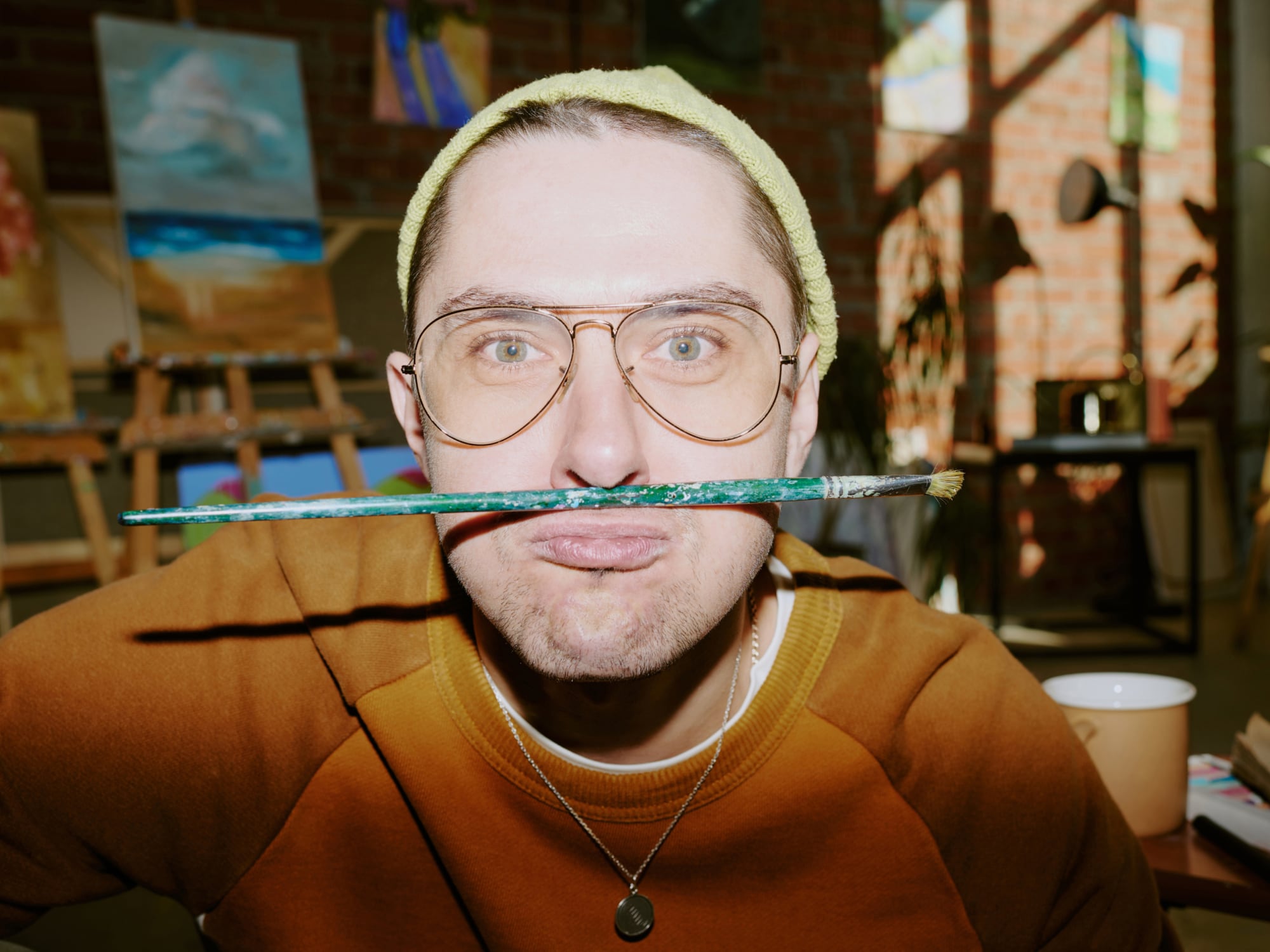 NDS-Kultusministerium_Erzieher-M1 Ein Mann mit heller Haut, Brille und grünem Beanie hält einen Pinsel horizontal unter seiner Nase und macht eine spielerische Grimasse. Er trägt ein braunes Sweatshirt und eine silberne Kette. Im Hintergrund steht eine Staffelei mit mehreren Leinwänden, die bunte Gemälde zeigen. Der Raum ist ein helles Atelier mit Backsteinwänden und großen Fenstern, durch die Sonnenlicht einfällt.