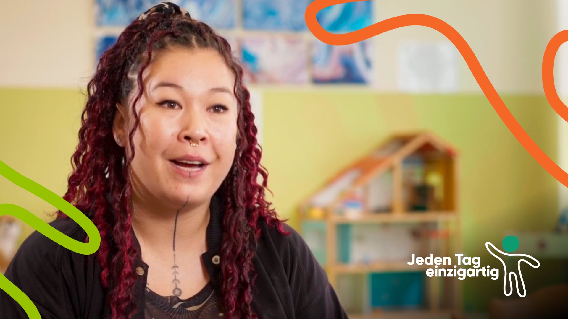 KM_Thumb-Video1 Eine Frau mit lockigen, dunkelroten Haaren, Gesichtspiercings und Tattoos spricht mit freundlichem Ausdruck. Im Hintergrund eine grüne Wand mit Kinderzeichnungen und ein Holzpuppenhaus. Orange und grüne grafische Elemente verlaufen durchs Bild. Logo 'Jeden Tag einzigartig' unten rechts.